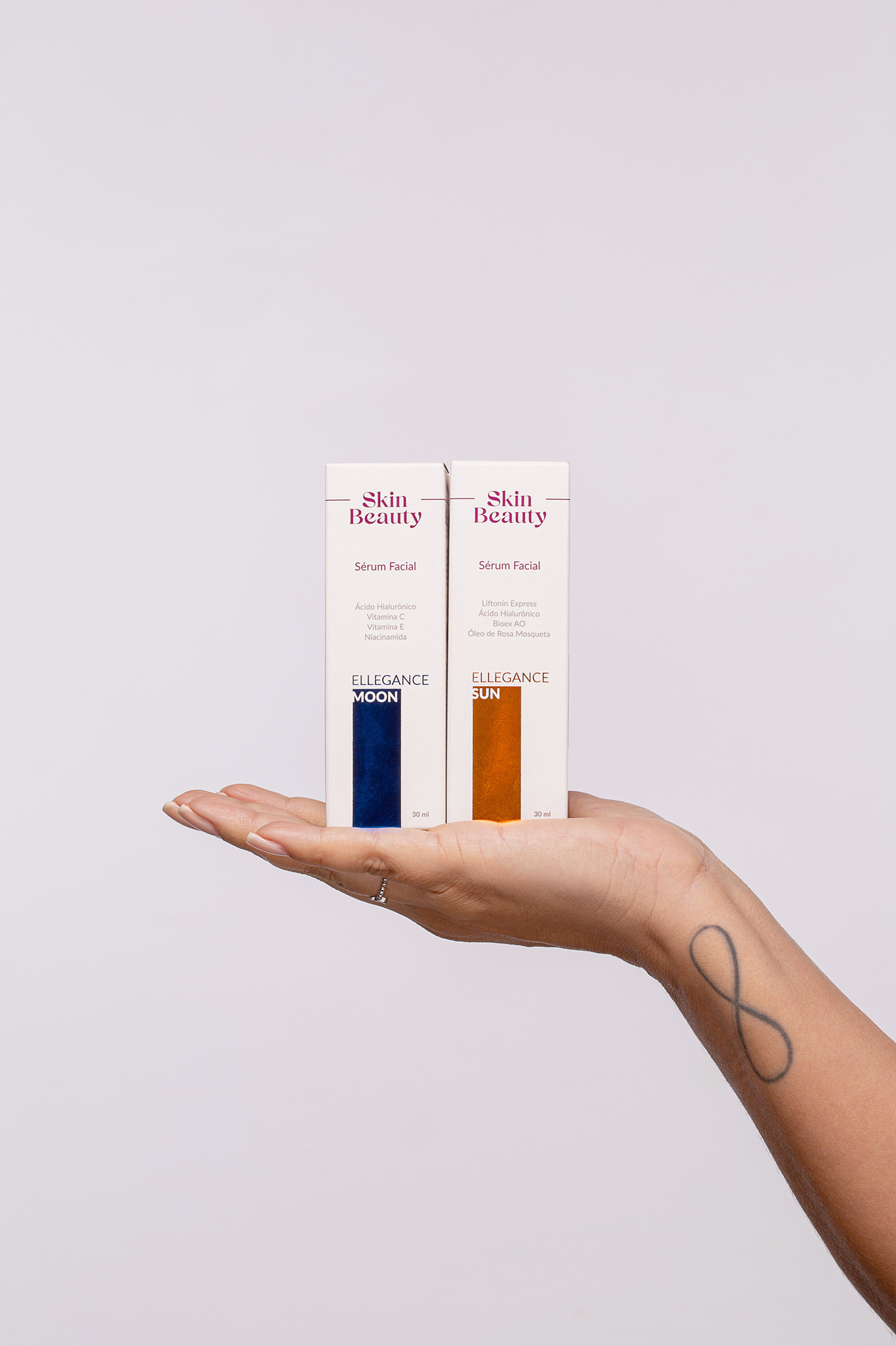 Duas embalagens do Sérum Facial Skin Beauty Ellegance — Moon e Sun — equilibradas na palma de uma mão, com um fundo neutro